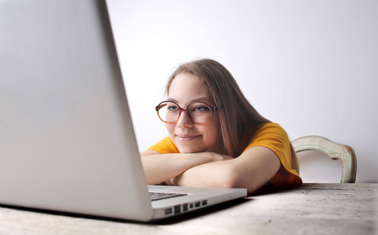 Young woman with glasses smiles while using a laptop at a desk indoors.