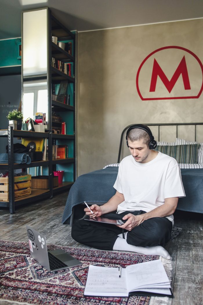 Adult man using a digital tablet with headphones in a modern bedroom setting, working remotely.
